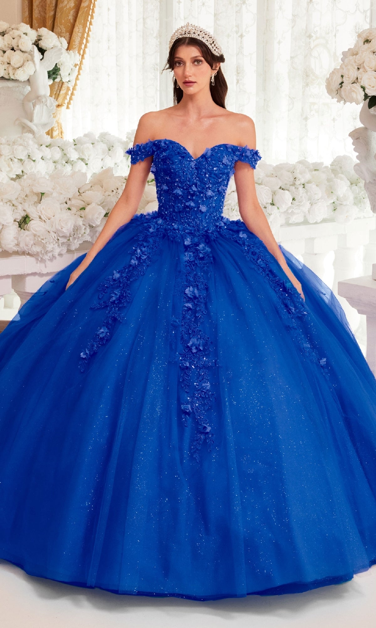 A woman wearing the Off-Shoulder Purple Quinceanera Dress, Ladivine 15717, stands indoors in front of white flowers and light curtains.